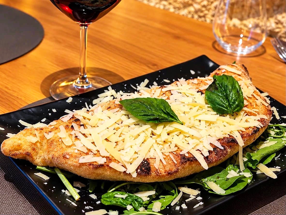 Il Calzone Classico su un piatto nero rettangolare: la sfoglia dorata coperta di scaglie di Parmigiano Reggiano con due foglie di basilico, su un letto di rucola, con un calice di rosso sul tavolo di rovere.