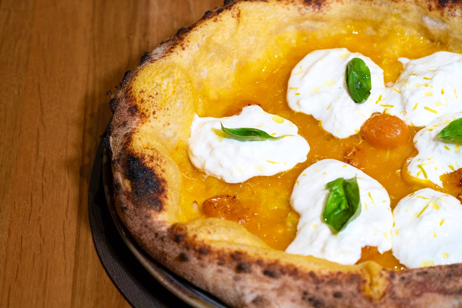 Primo piano della Cortile Chiaramonte: salsa di datterino giallo, grandi ciuffi di stracciatella, datterini gialli confit, foglie di basilico e scorza di limone grattugiata, su un cornicione alto e dorato, sul tavolo di rovere.