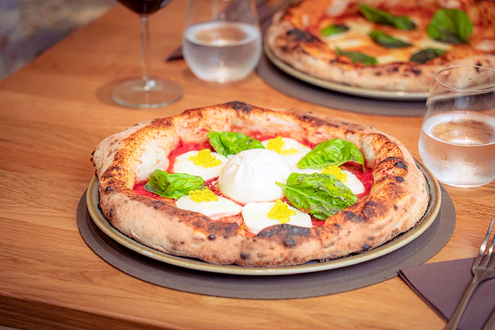 Un tavolo di rovere apparecchiato: la margherita contemporanea con una burrata intera al centro, le perle gialle di olio extravergine e il basilico, con i calici, la caraffa d'acqua e una seconda pizza dietro.