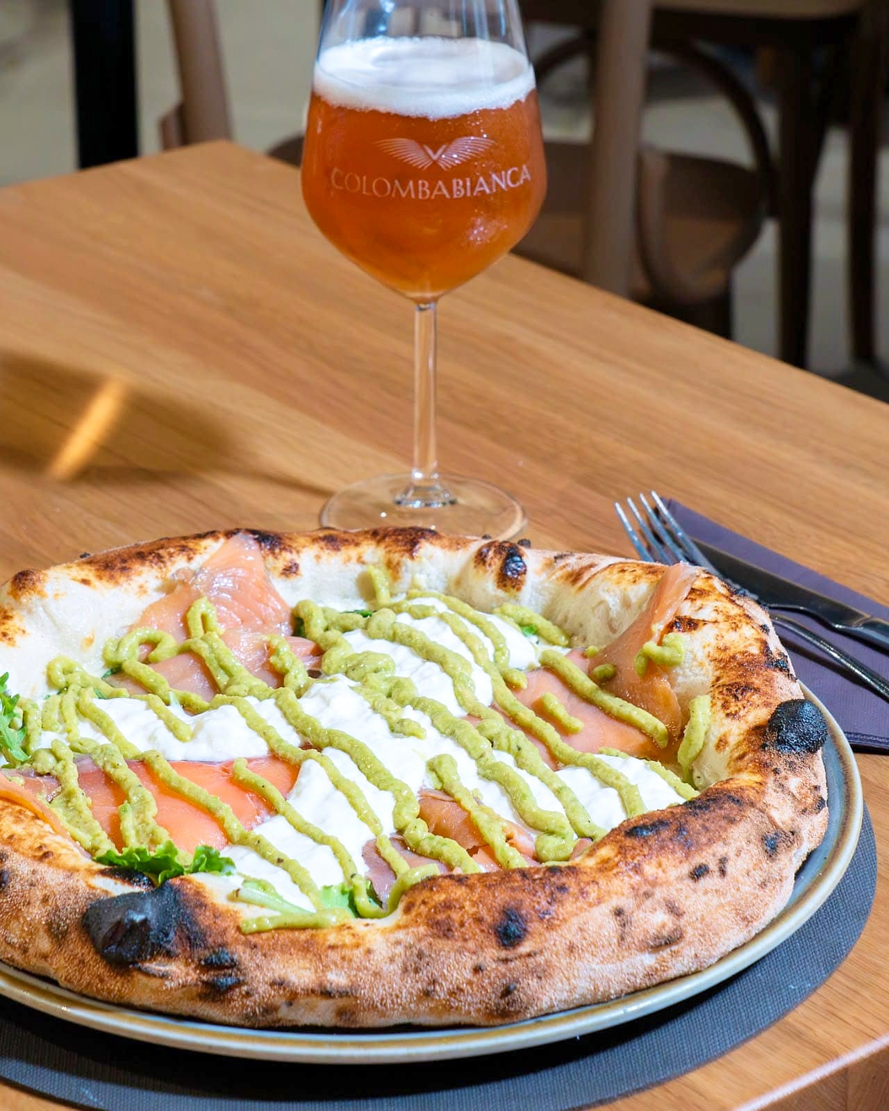 Pizza con salmone affumicato, stracciatella e strisce di guacamole su un cornicione alto e maculato dalla cottura, accanto a un calice di birra.