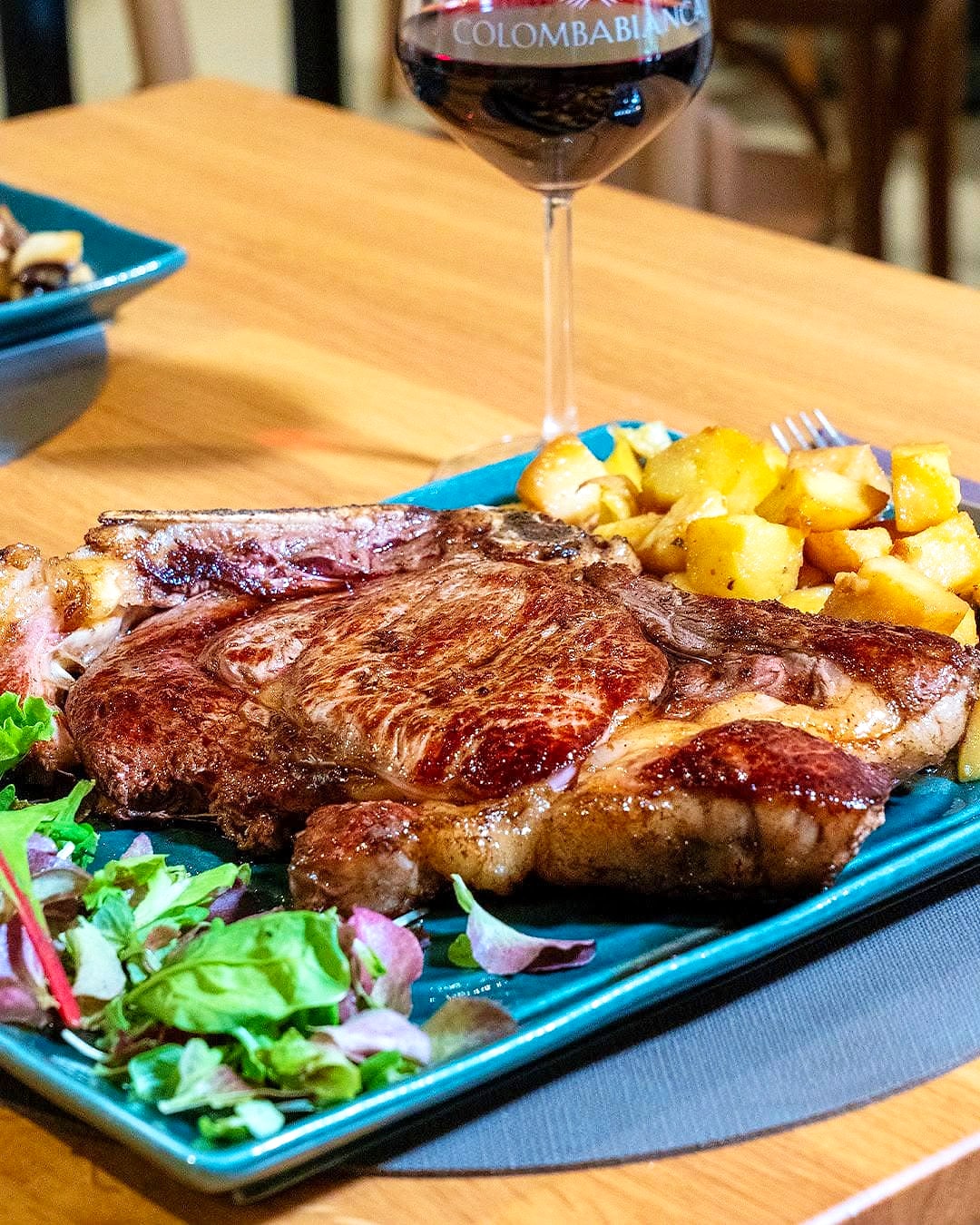 Fiorentina alla griglia su un piatto di ceramica verde petrolio, con patate al forno e insalata, e un calice di vino rosso sul tavolo di legno.