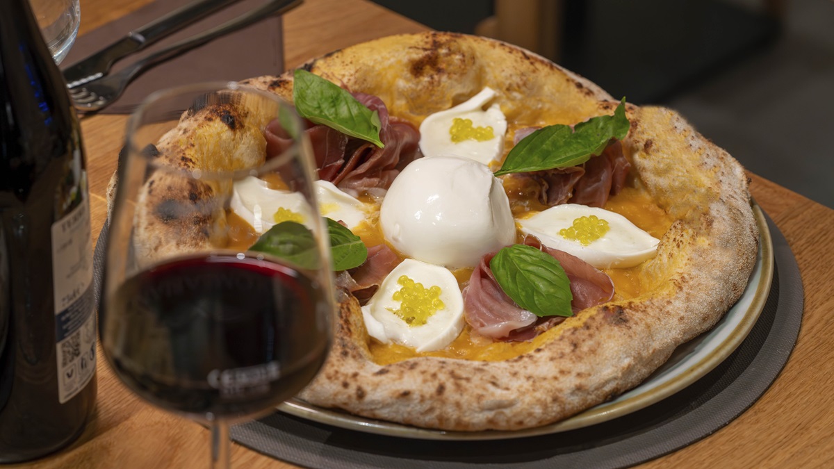 Pizza con salsa di datterino giallo, mozzarella, prosciutto crudo, basilico e perle verdi di olio extravergine, accanto a un calice di vino rosso.