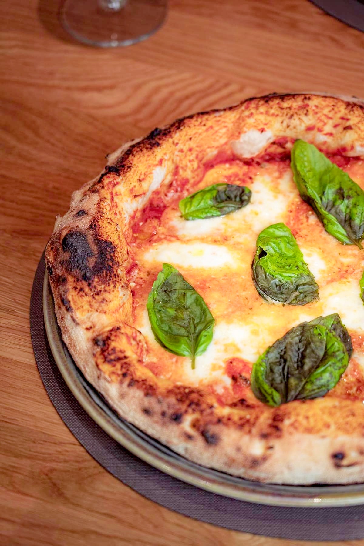 La Margherita appena uscita dal forno: il pomodoro, il fiordilatte sciolto a chiazze, le foglie di basilico e un filo d'olio, su un cornicione alto e bruciacchiato dalla fiamma, sul tavolo di rovere.