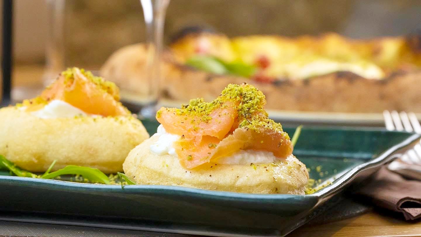 Due montanarine fritte su un piatto rettangolare di ceramica verde petrolio, con la stracciatella, il salmone affumicato e la polvere di pistacchio.