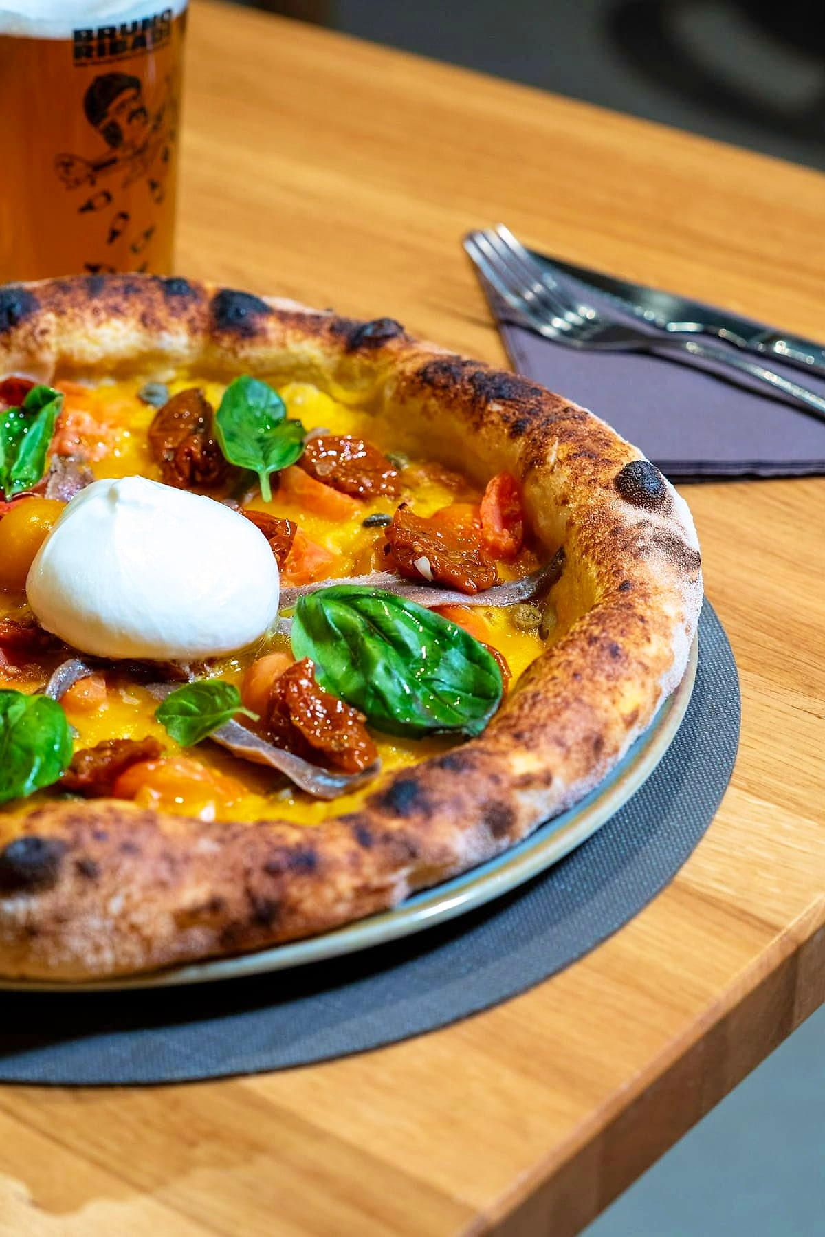 Pizza su base di datterino giallo con una burrata intera al centro, alici di Cetara, pomodorini essiccati, capperi e foglie di basilico, accanto a un boccale di birra artigianale.