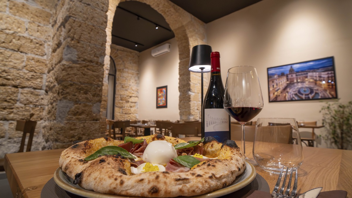 La sala la sera vista da un tavolo: l'arco di tufo, una lampada nera, una bottiglia di vino rosso e una pizza in primo piano.
