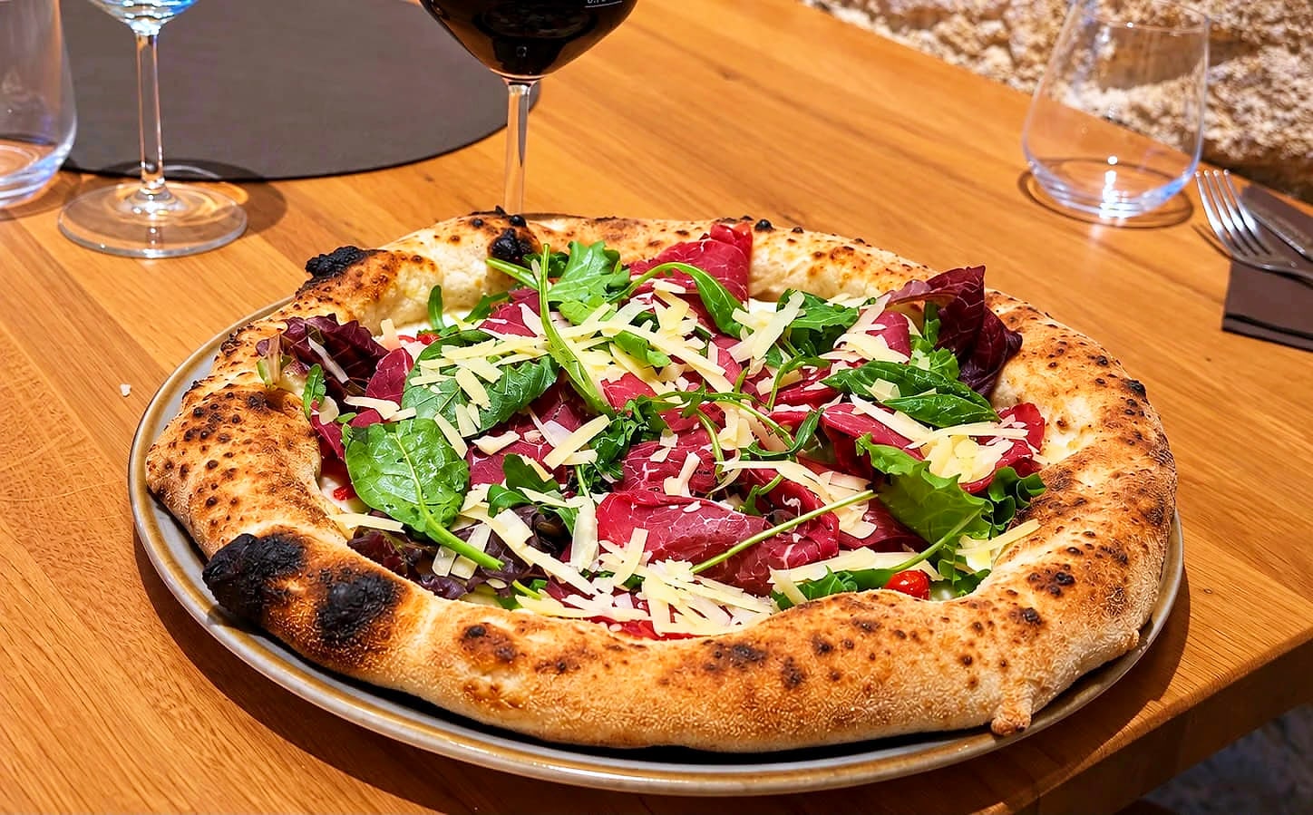 La Cortile San Bonomo sul tavolo di rovere: fette di carpaccio di manzo, rucola e insalatina, scaglie di Parmigiano Reggiano e datterini rossi su un cornicione alto e maculato dalla cottura, con un calice di rosso dietro e il muro di tufo.