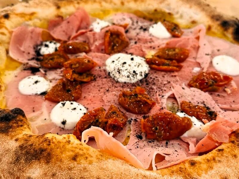 La Cortile Sclafani vista dall'alto: prosciutto cotto affumicato, ciuffi di ricotta fresca, datterini rossi confit e polvere di olive nere su un cornicione dorato.