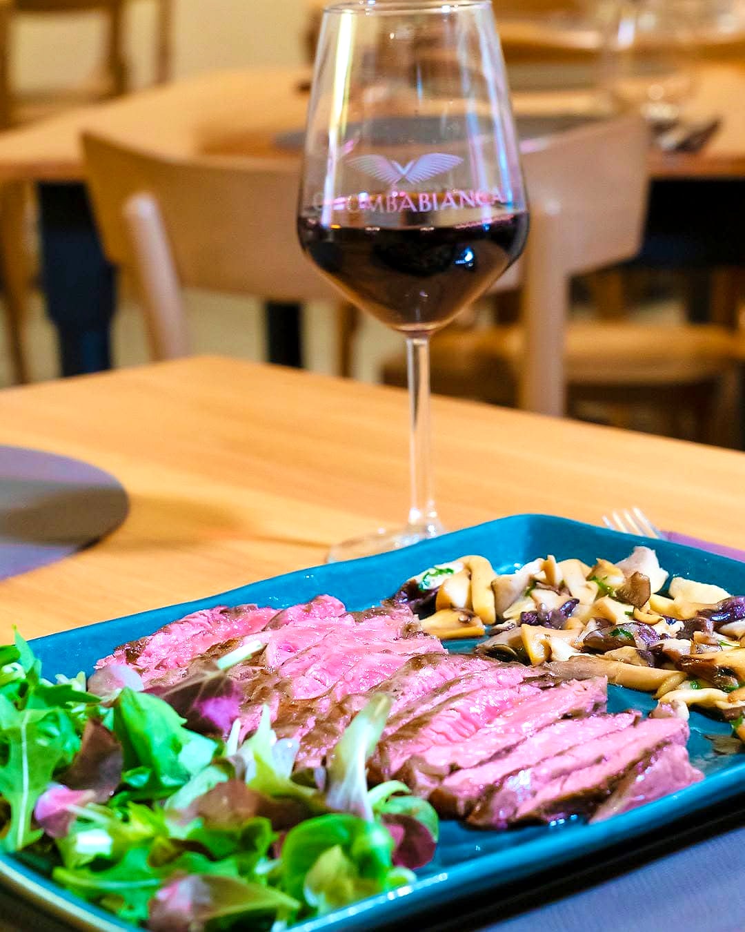 Tagliata di Angus al sangue affettata su un piatto rettangolare di ceramica verde petrolio, con i funghi cardoncelli saltati e l'insalatina, e un calice di vino rosso sul tavolo di rovere.