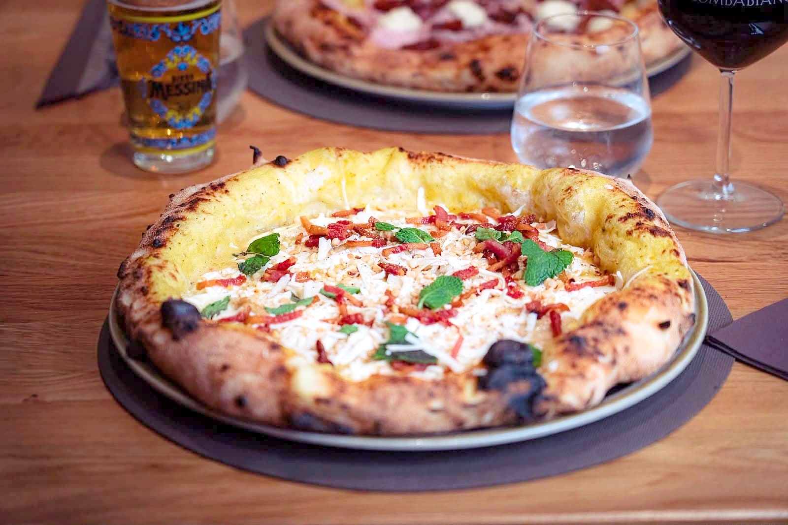 Un tavolo di rovere la sera: un bicchiere di birra, un calice di vino rosso e un bicchiere d'acqua intorno a una pizza dal cornicione alto e dorato, con una seconda pizza sullo sfondo.