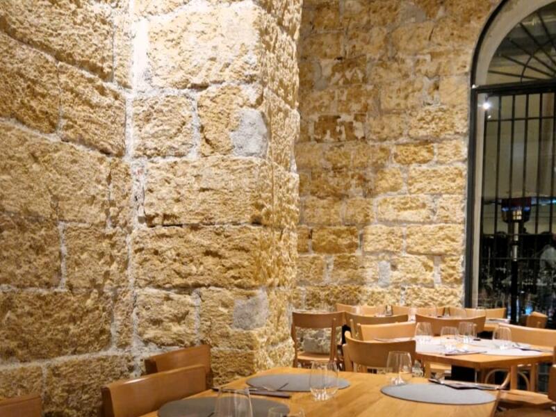 Un pilastro di conci di tufo con i giunti di calce bianca, illuminato dal basso, e i tavoli di frassino della sala.