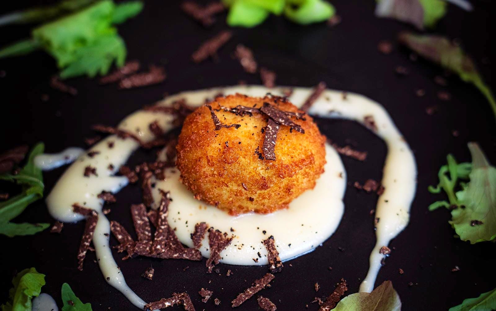 Uovo in mollica croccante, dorato, al centro di una fonduta di Parmigiano su un piatto nero, con scaglie di tartufo nero tutt'intorno e foglie di insalatina.