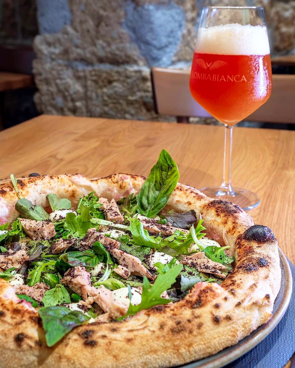Pizza con filetti di tonno, insalata mista, ciuffi di salsa tonnata e una spolverata scura, davanti a un calice di birra rossa e al muro di tufo della sala.
